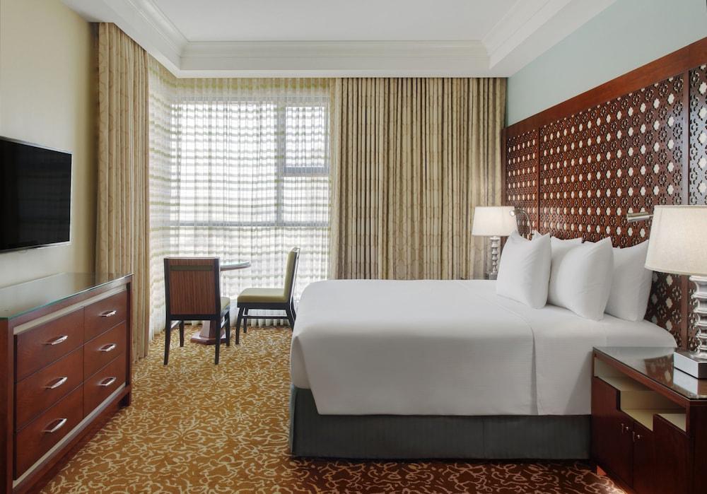 Hilton Suites Jabal Omar Makkah Mekka Exteriér fotografie The photo shows a hotel room featuring a large bed with white linens and several pillows. There is a wooden headboard with a decorative design. Alongside the bed, there are bedside lamps. A wooden dresser is visible, with a television mounted on the 