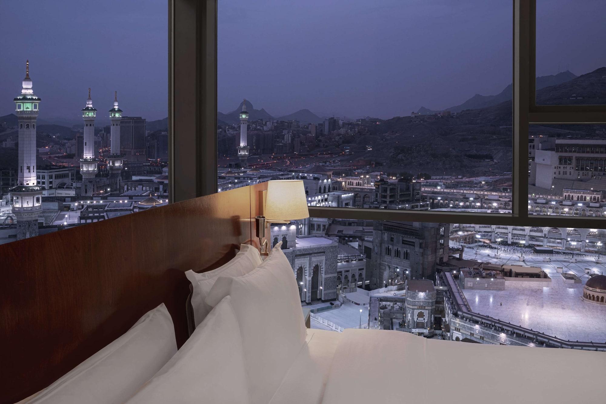 Hilton Suites Jabal Omar Makkah Mekka Exteriér fotografie The photo depicts a view from inside a hotel room at night. The foreground shows a bed with white pillows and linens. In the background, large windows offer a panoramic view of a cityscape, featuring notable minarets and buildings illuminated against