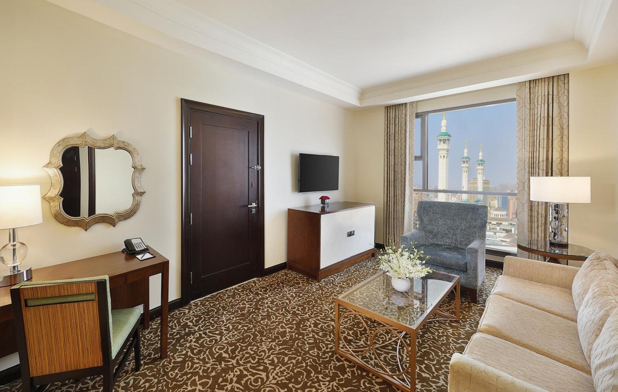 Hilton Suites Jabal Omar Makkah Mekka Exteriér fotografie The photo shows a hotel room with a modern and elegant design. It features a cozy seating area with a beige sofa and a blue armchair, complemented by a glass coffee table. There is a wooden desk with a mirror and a phone, and a television mounted on 