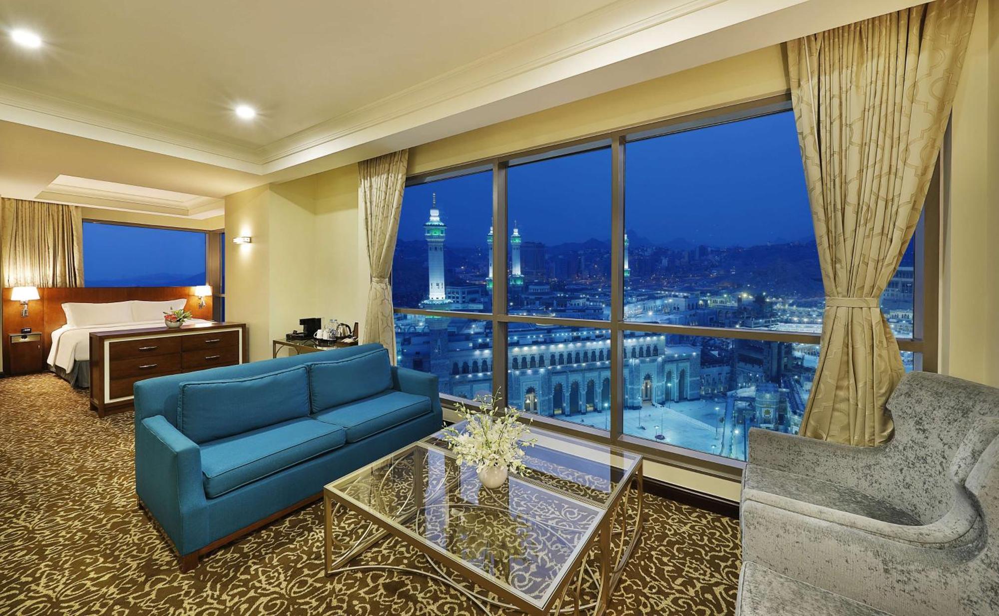 Hilton Suites Jabal Omar Makkah Mekka Exteriér fotografie The photo shows a luxurious hotel room with a stunning view. The room features a modern design, including a blue sofa and a glass coffee table on a patterned carpet. Large windows provide an expansive view of a cityscape, illuminated at night, showca
