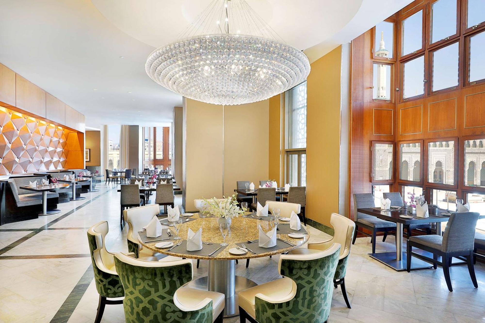 Hilton Suites Jabal Omar Makkah Mekka Exteriér fotografie The photo shows a modern restaurant interior. In the center, there's a round dining table with green upholstered chairs, elegantly arranged with plates and napkins. Above the table hangs a large crystal chandelier, providing bright, stylish lighting.