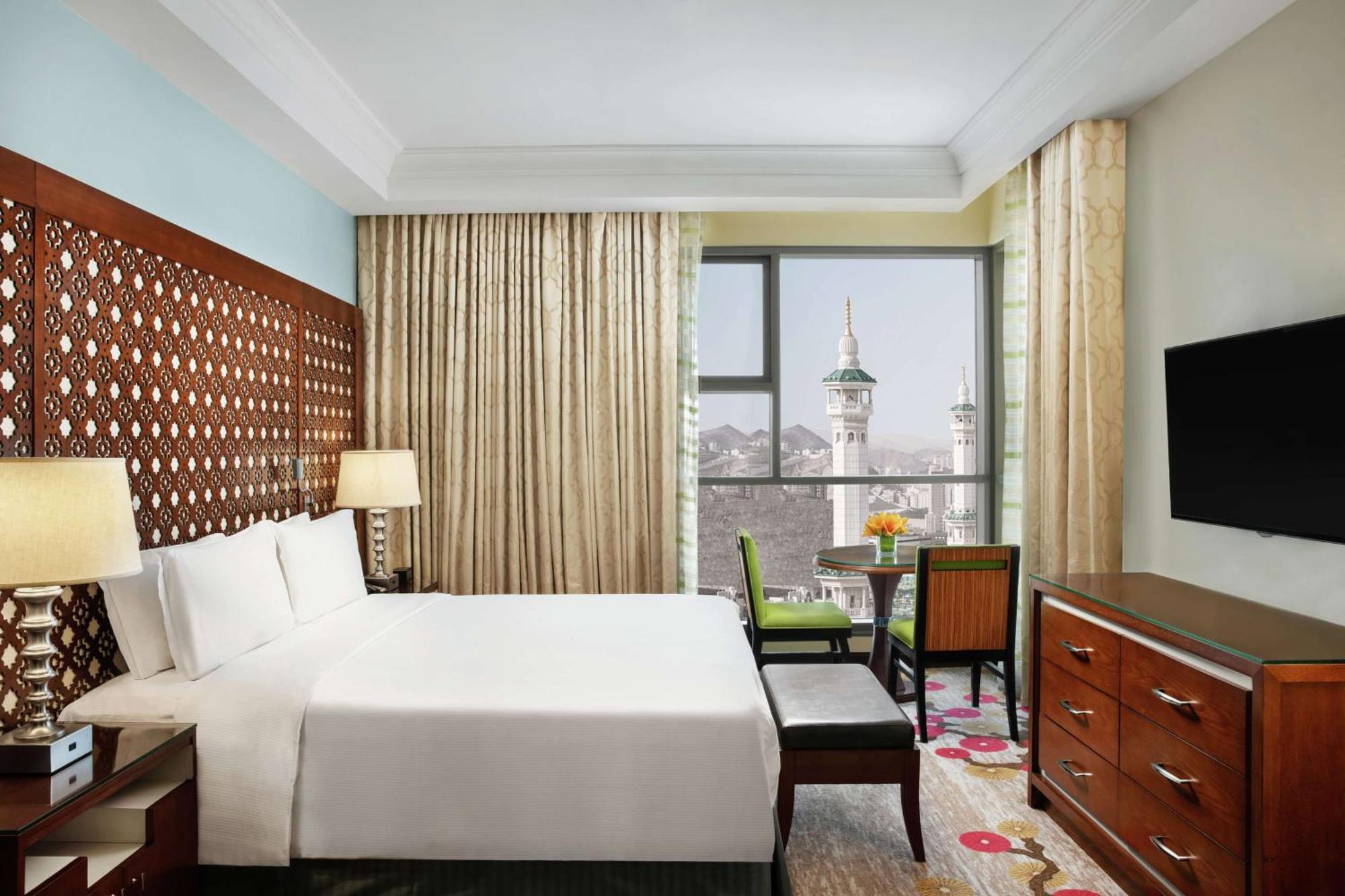 Hilton Suites Jabal Omar Makkah Mekka Exteriér fotografie The photo shows a well-furnished hotel room. It features a large bed with white bedding, a wooden headboard, and two bedside lamps. There’s a small seating area with a chair and a table near a window that provides a view of a mosque's minarets outsid