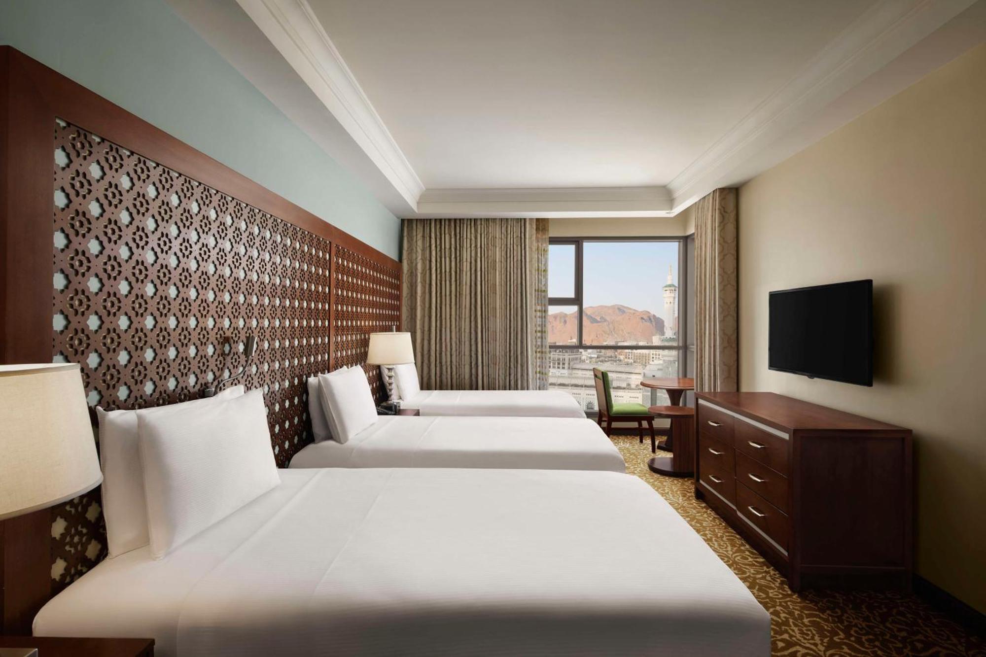 Hilton Suites Jabal Omar Makkah Mekka Exteriér fotografie The photo shows a spacious hotel room featuring three single beds arranged in a row. Each bed has white linens and a headboard with a decorative design. There's a large window that lets in natural light, with curtains drawn to the side, revealing a v