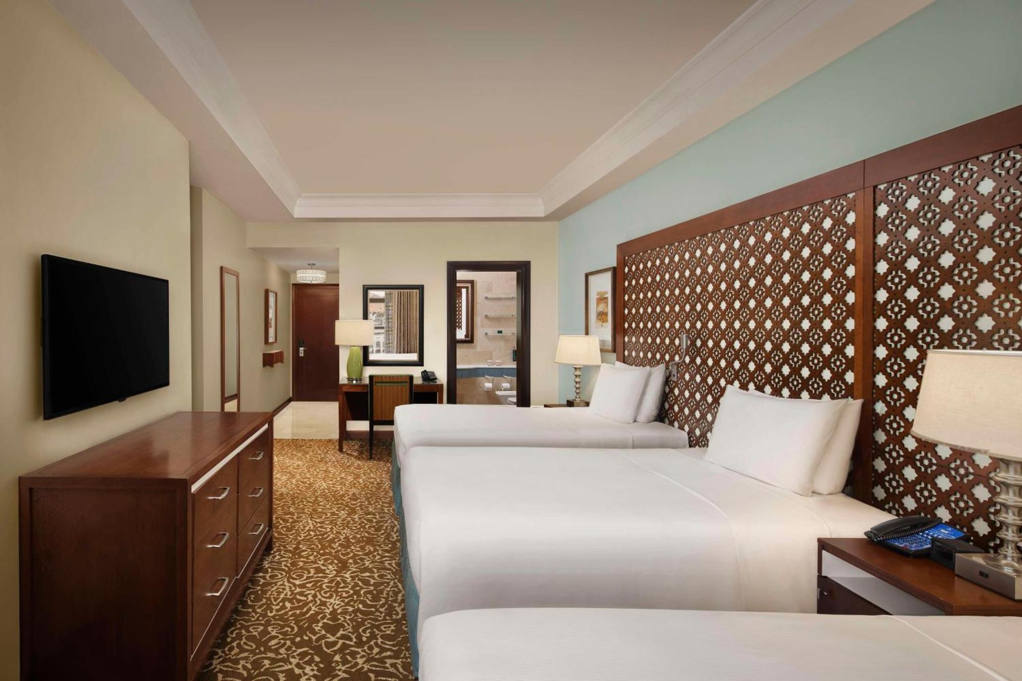 Hilton Suites Jabal Omar Makkah Mekka Exteriér fotografie The photo shows a hotel room featuring two beds with white linens. There is a decorative headboard behind each bed, made of wood with a patterned design. The room has a warm beige carpet and light blue walls, giving it a serene atmosphere. A wooden d