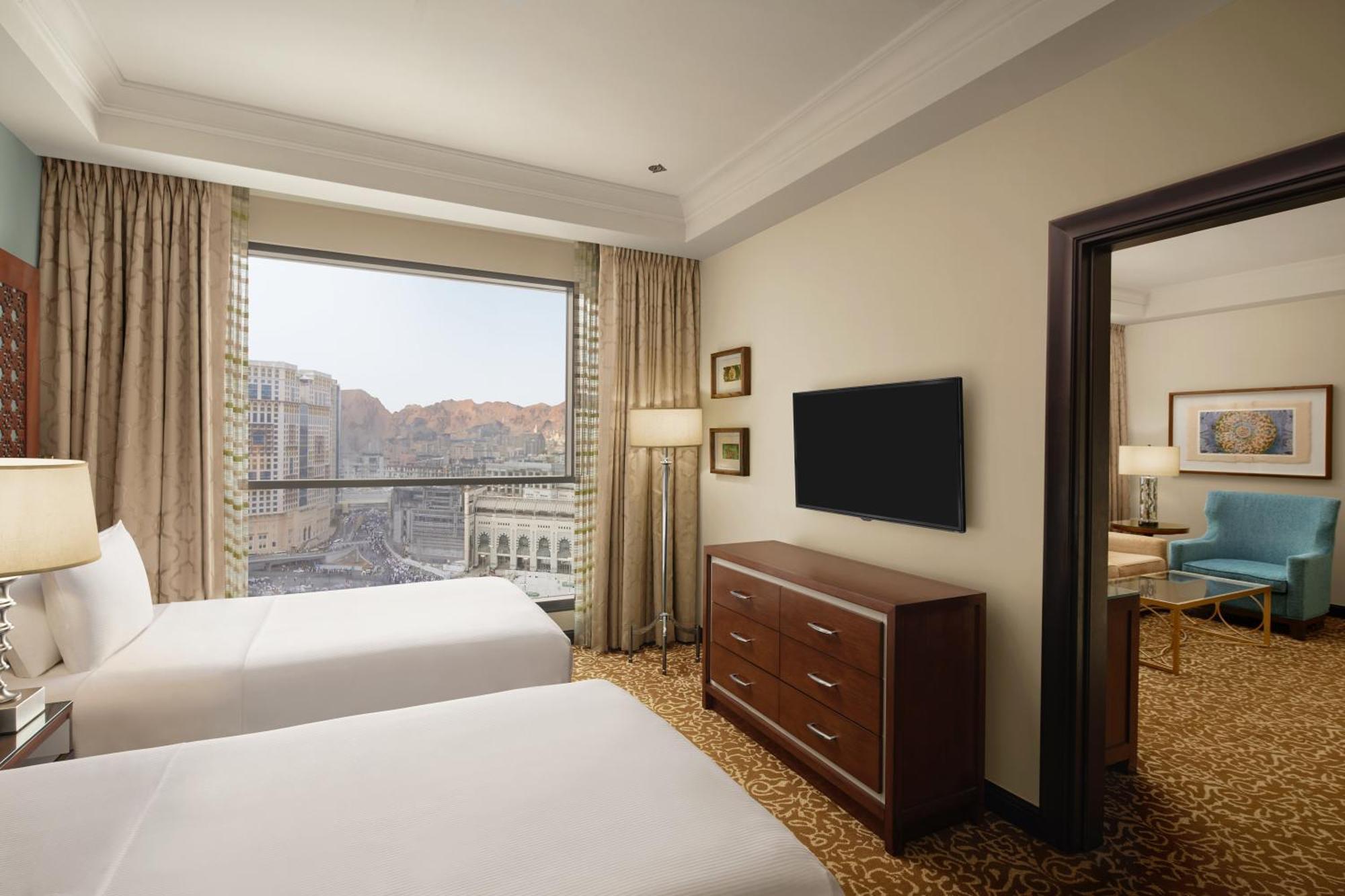 Hilton Suites Jabal Omar Makkah Mekka Exteriér fotografie The photo shows a hotel room featuring two beds with white bedding. There is a large window with curtains that allow for natural light, offering a view of the outside scenery, which includes mountains. In the room, there is a wooden dresser and a fla
